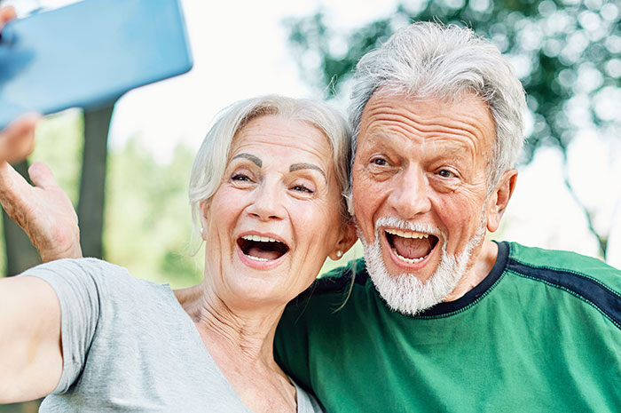 Happy elderly couple taking a selfie outdoors, capturing the surprise moment of husband&rsquo;s parents visiting on honeymoon trip.