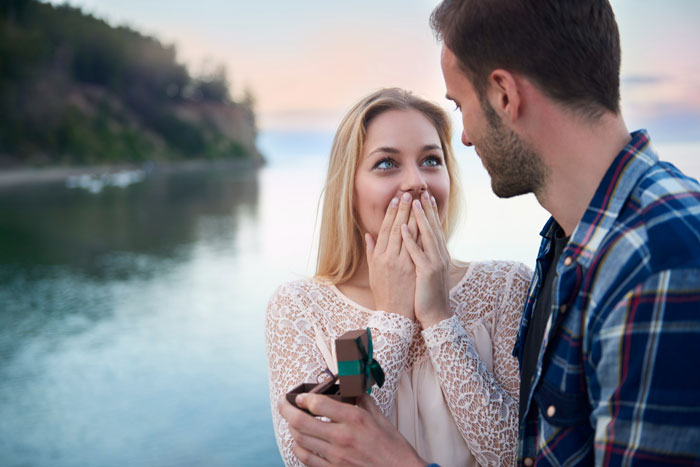 Couple by the water with man proposing and woman reacting with surprise, illustrating a Venmo search unraveling a fabricated past. Couple by the water with man proposing and woman reacting with surprise, illustrating a Venmo search unraveling a fabricated past.