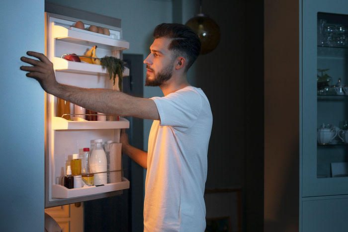 Man standing by an open fridge at night, reflecting on a moment after embarrassing wife in front of her parents.