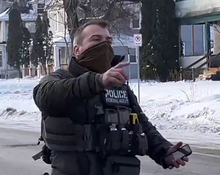 ICE agent in tactical gear and face mask holding phone, standing outside in snowy residential neighborhood.