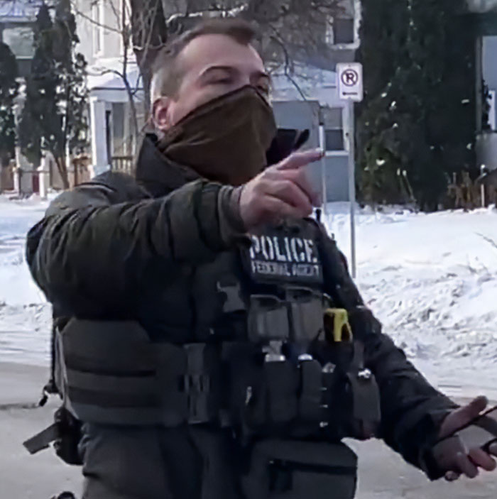 Minneapolis ICE agent wearing tactical gear and face covering gestures while speaking outdoors in snowy neighborhood.