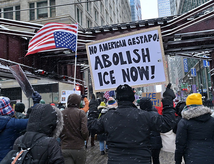 Protesters in winter coats hold signs calling to abolish ICE during a heated demonstration on a snowy city street.