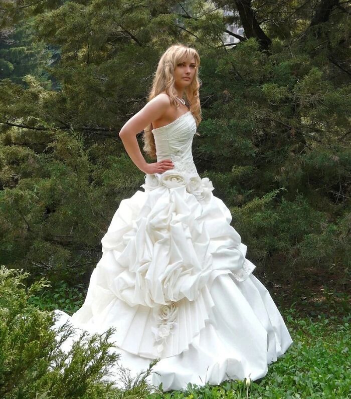 Woman posing outdoors in a white wedding dress with elaborate ruffles and floral details, showcasing questionable taste wedding dresses.