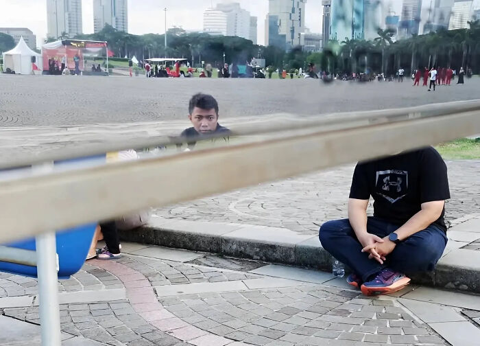 Man sitting on pavement partially hidden behind a horizontal railing in a perfectly timed street shot by a photographer.