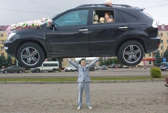 Man in a suit seemingly lifting a car with a bride inside, an awkward wedding photo capturing a funny moment.