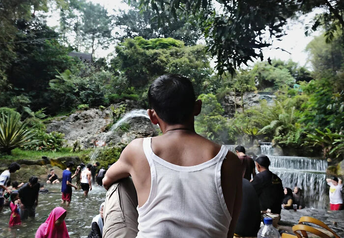 Man in white tank top sitting by a forest stream with people enjoying water in the background street shots perfectly timed