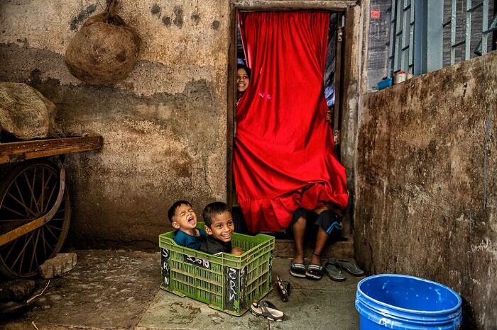Two children laugh playing inside a green crate near an old wall, while a woman peeks from behind a red curtain in a candid street photo.
