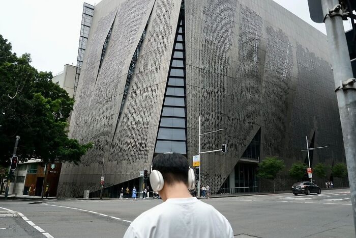 Person wearing headphones standing at a street corner with modern building, capturing perfect coincidence moment in street photography.