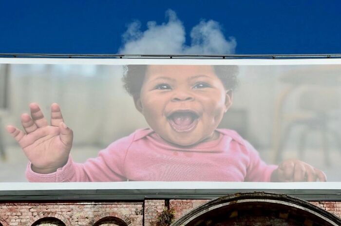 Smiling baby on billboard perfectly aligned with smoke cloud, showcasing street photographer's talent for capturing perfect coincidences.