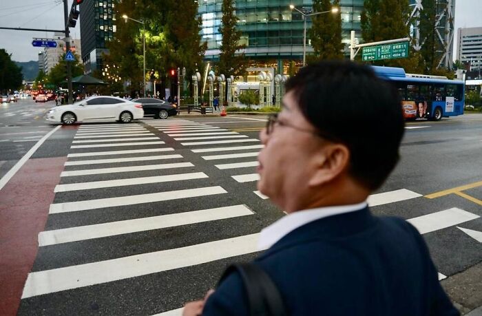 Man standing at busy urban crosswalk with cars and bus, captured by street photographer skilled in perfect coincidences.