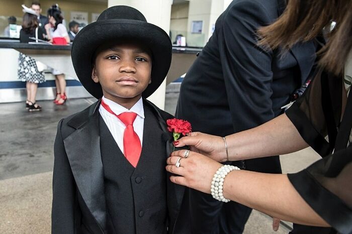 Young boy dressed in a suit and hat having a red flower pinned, candid street photo capturing tender human moments.