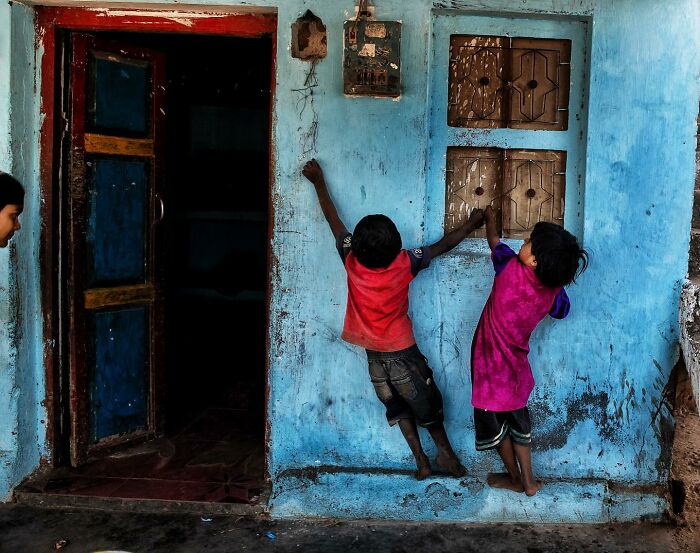 Two children playing and climbing on a weathered blue wall in a candid street photo capturing humanity and tender moments.