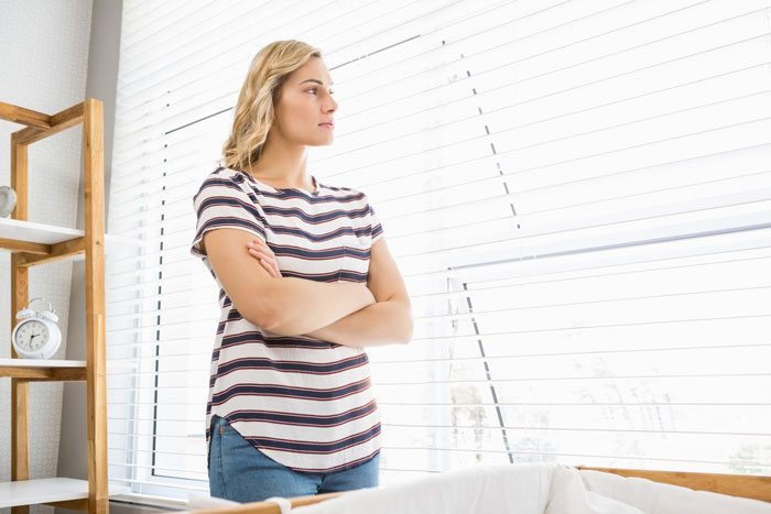 Woman standing with arms crossed by window blinds, reflecting tension in a hostage dinner and car food threat scenario.
