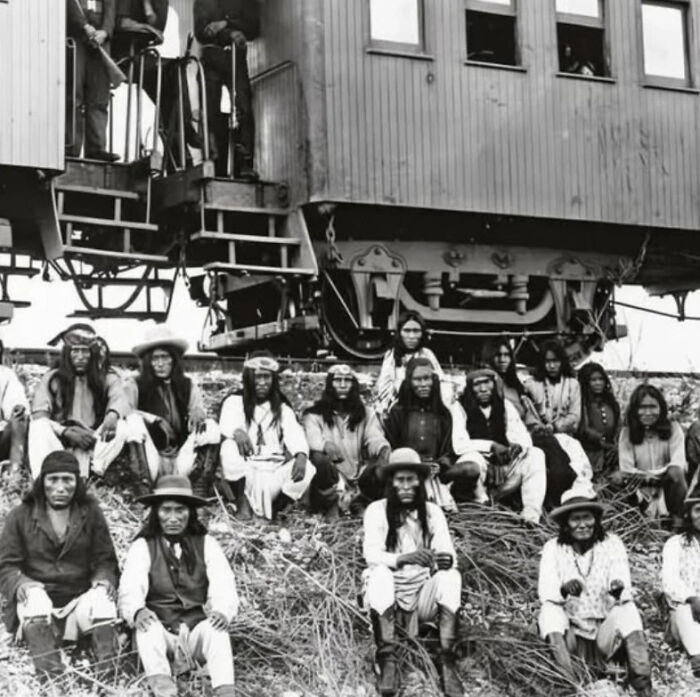 Group of indigenous men seated near a train car, a powerful photograph reflecting the history of humanity.