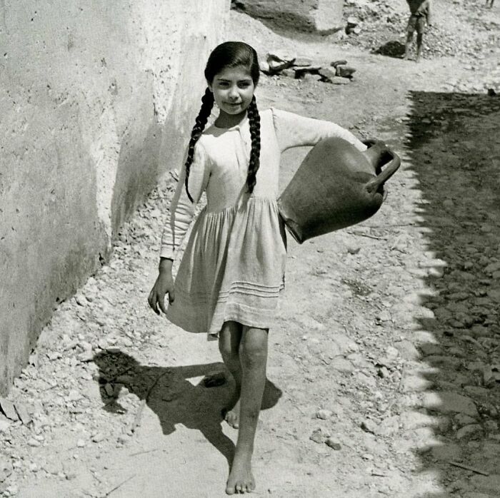 Young girl with braided hair carrying a large container barefoot on a dirt path in a historic photograph of humanity.