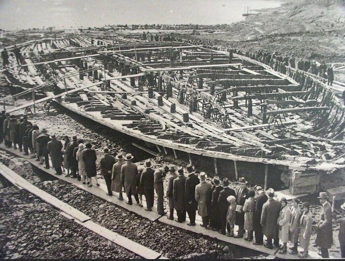 Large group of people observing the construction of an ancient shipwreck during a historical excavation.