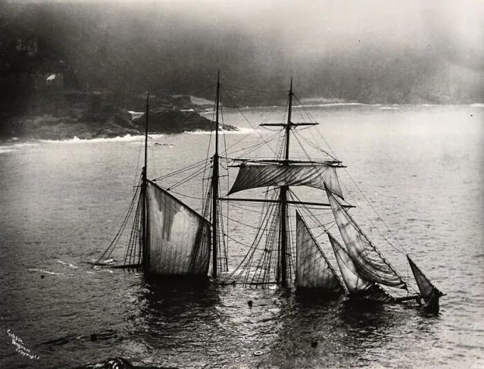 Sinking sailing ship with torn sails in water near rocky shore, capturing a tragic moment in the history of humanity.