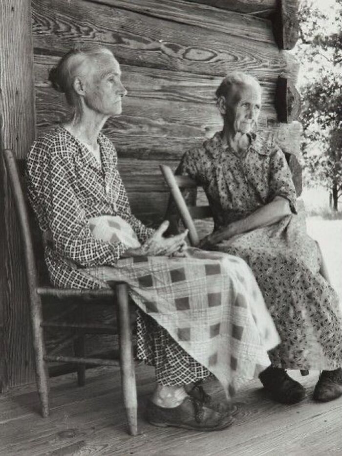 Two elderly women sitting on wooden chairs outside a rustic cabin, depicting the sad and beautiful history of humanity.
