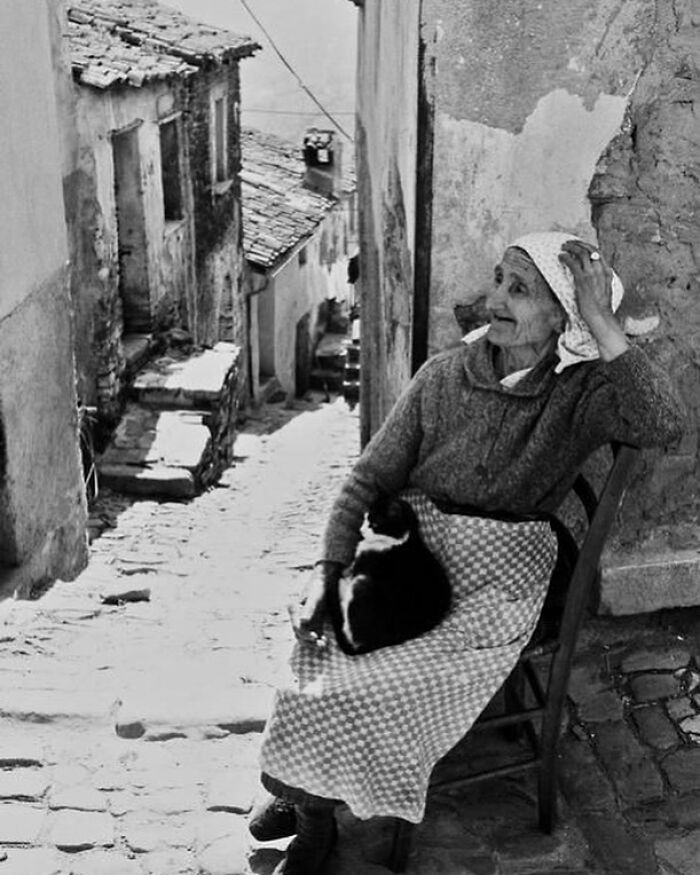 Elderly woman sitting on chair in narrow old street with cat on her lap, reflecting the history of humanity.