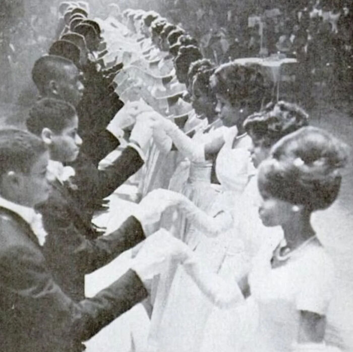 Black and white historic photograph showing young couples in formal attire holding hands during a ballroom dance event.