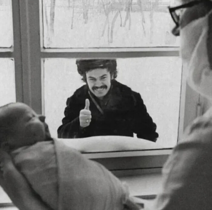 Man outside window giving thumbs up while a medical professional holds a newborn baby showing history of humanity moments.