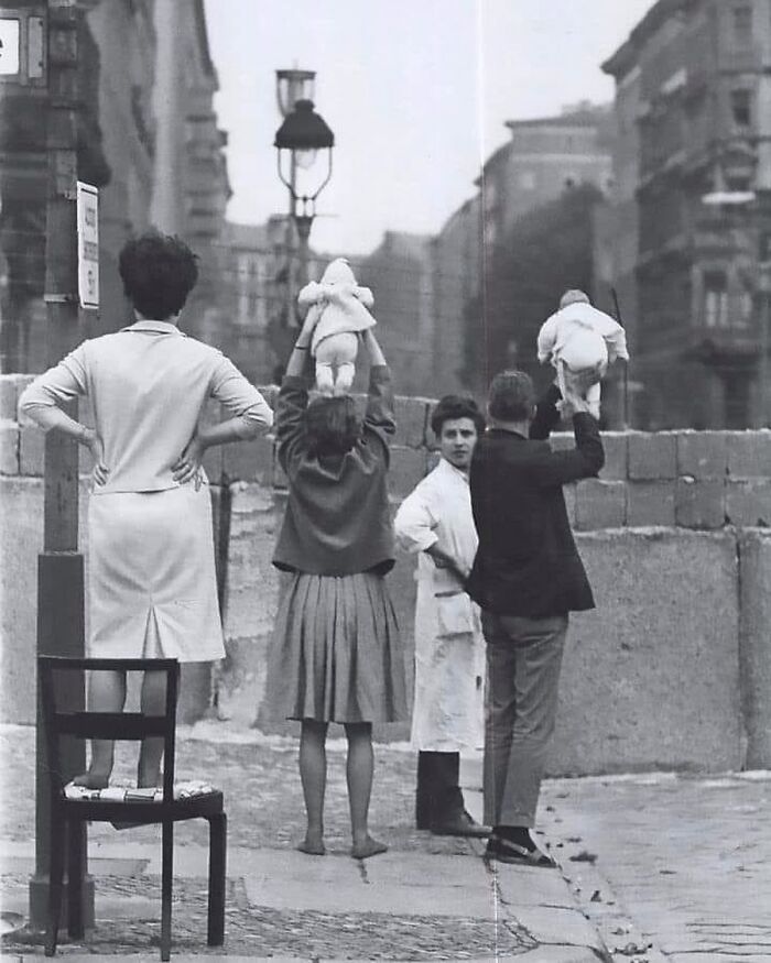 Black and white photograph showing people holding babies over a wall, symbolizing the complex history of humanity.