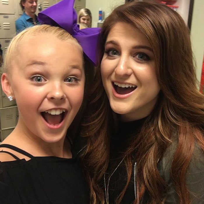 JoJo Siwa with a large purple bow and another woman smiling excitedly in a casual indoor setting. JoJo Siwa with a large purple bow and another woman smiling excitedly in a casual indoor setting.