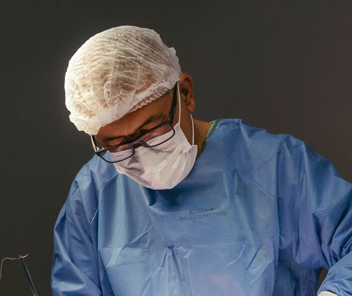 Doctor wearing surgical cap and mask, focusing intently while performing a medical procedure on a patient in a clinical setting.