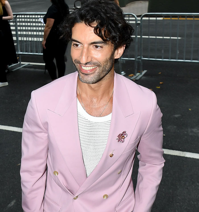 Justin Baldoni smiling in a light pink blazer with a white mesh shirt at an outdoor event with metal barriers behind him