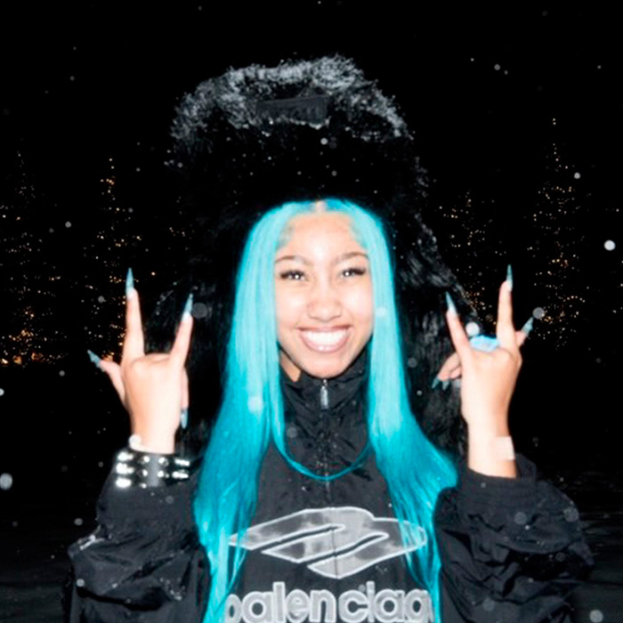 Young woman with bright blue hair and black fur hat smiling and making hand signs at night outdoors.