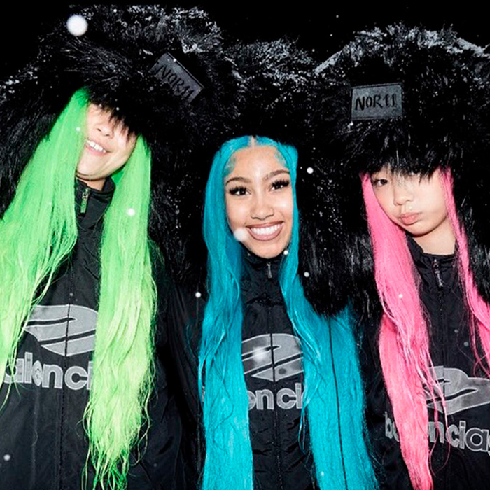 Three women wearing colorful wigs and black fur hats, styled fashionably amid winter snowfall, in a bold group look.