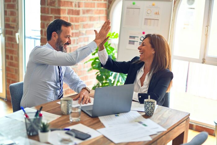Two colleagues in business attire high-fiving over a desk in an office, illustrating overrated adult goals.