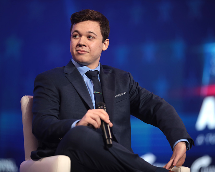 Kyle Rittenhouse in a dark suit holding a microphone, reacting during a public event amid internet controversy.