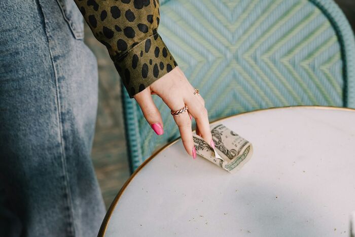 Woman with leopard print blouse and pink nails tipping a dollar on a table, illustrating things normal in America but offensive elsewhere.
