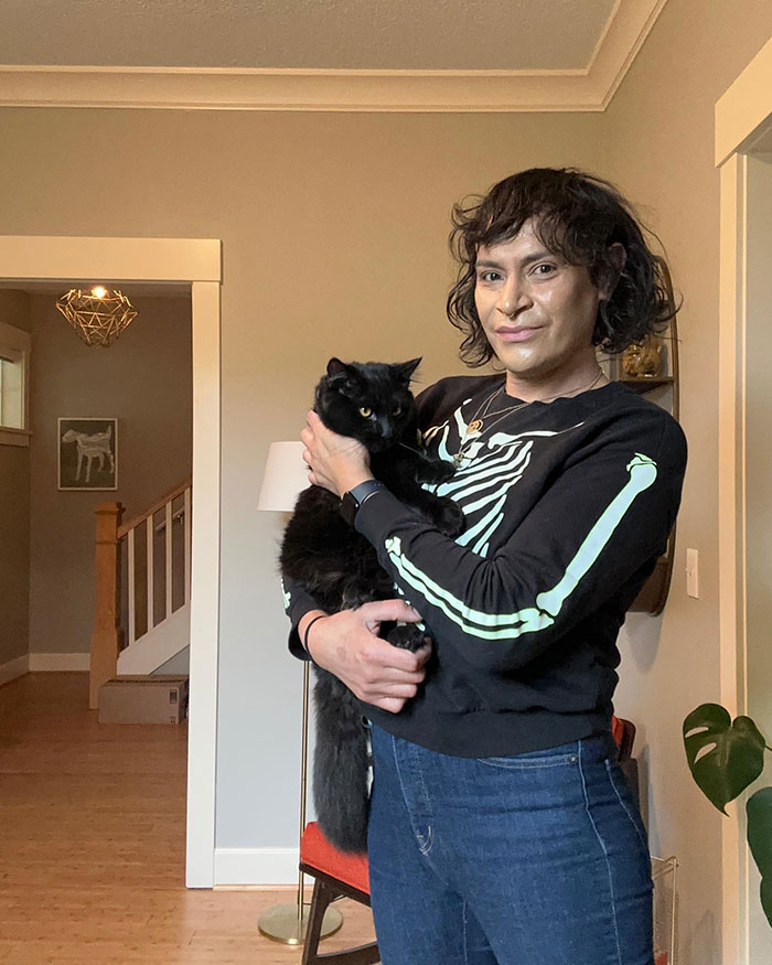 Person wearing a skeleton print sweater holding a black cat indoors, related to trans chef of the year media coverage. Person wearing a skeleton print sweater holding a black cat indoors, related to trans chef of the year media coverage.