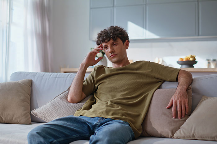 Young man sitting on sofa looking upset, reflecting on family forgiveness and conflicted feelings about their tone.