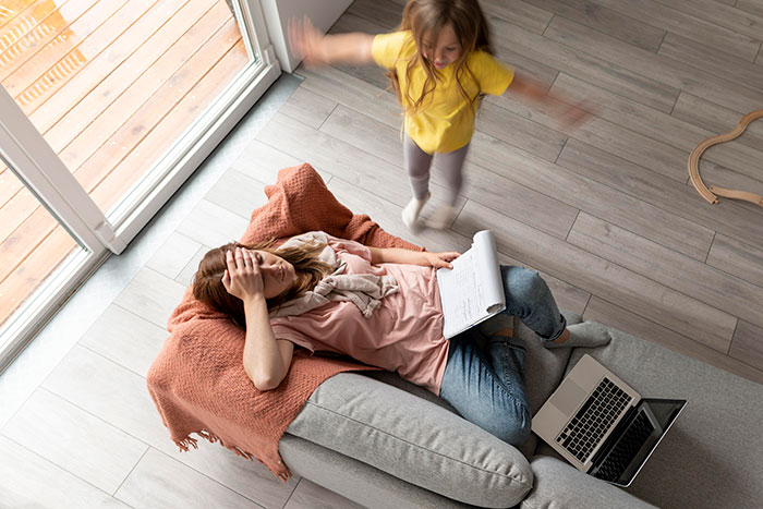 Stressed woman on couch with laptop and notebook as child jumps nearby, illustrating girlfriend ATM pressure Stressed woman on couch with laptop and notebook as child jumps nearby, illustrating girlfriend ATM pressure