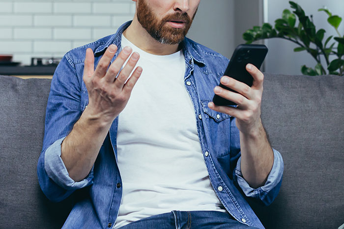Man sitting on couch looking shocked and confused while reading a message on his smartphone about wife’s cheating.