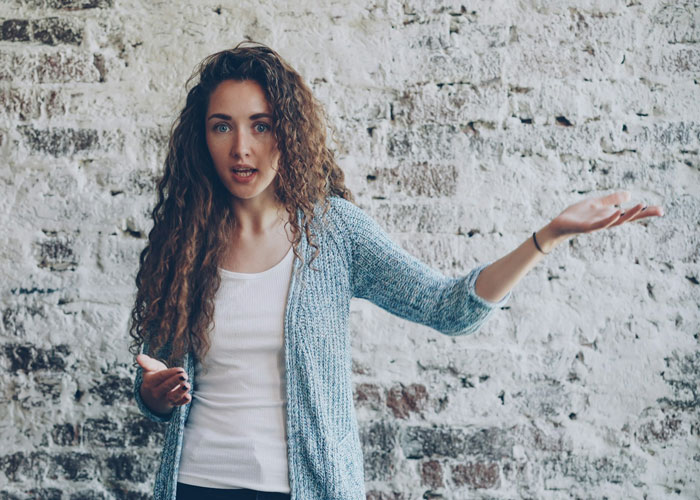 Unemployed woman with curly hair in casual clothes expressing frustration against a brick wall background.