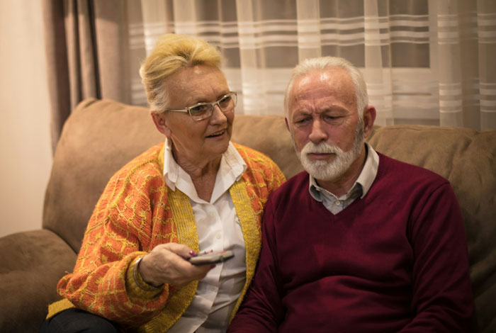 Elderly couple sitting on a couch, woman holding remote, reflecting tension with in-laws refusing to help with childcare.