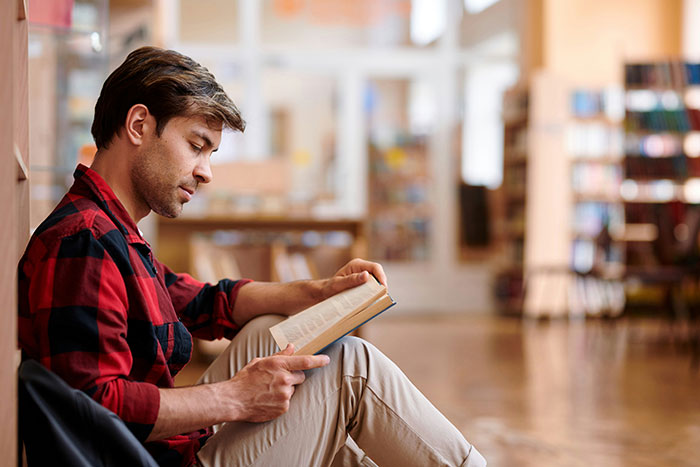 Man reading a book indoors, reflecting on revenge and reckoning against a coworker who got him fired.