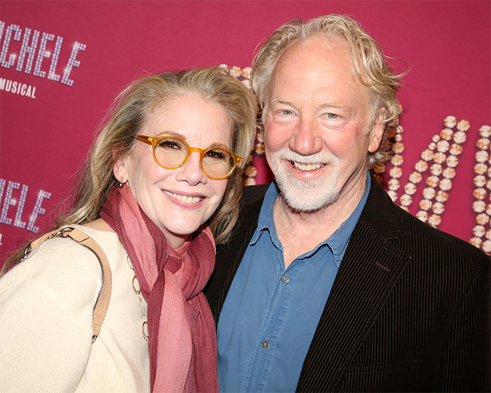 Melissa Gilbert and Timothy Busfield smiling together at a public event with a red backdrop behind them.