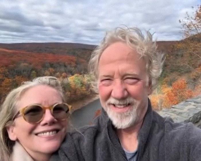 Melissa Gilbert and Timothy Busfield smiling outdoors with autumn foliage in the background during a scenic moment.