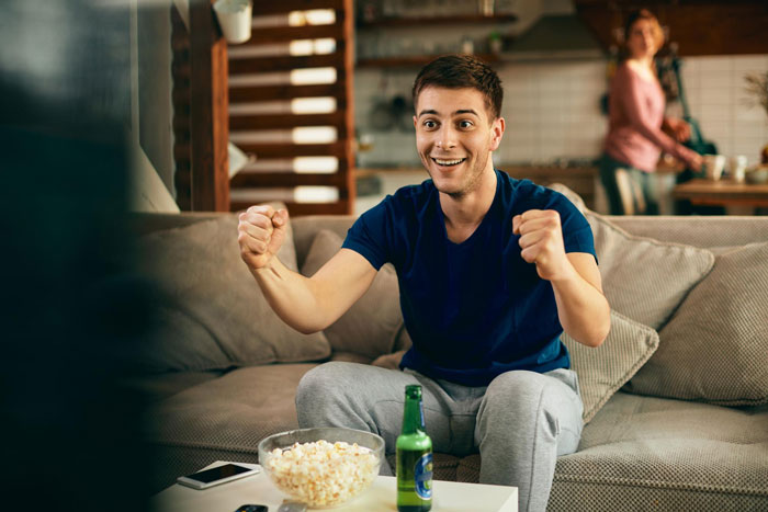 Man sitting on couch shows excited behavior, while a woman stands in the background in a home setting.