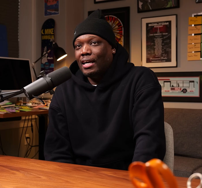 Michael Che wearing black hoodie and beanie speaking into a microphone during a podcast about SNL and ICE debate.