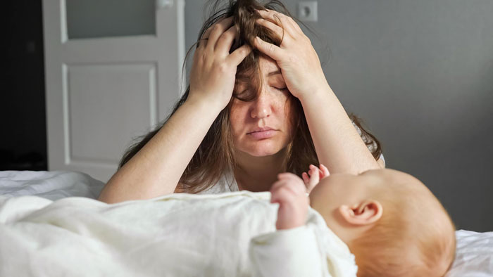 Tired mother resting head in hands while looking at newborn baby lying on bed in a quiet room. Tired mother resting head in hands while looking at newborn baby lying on bed in a quiet room.
