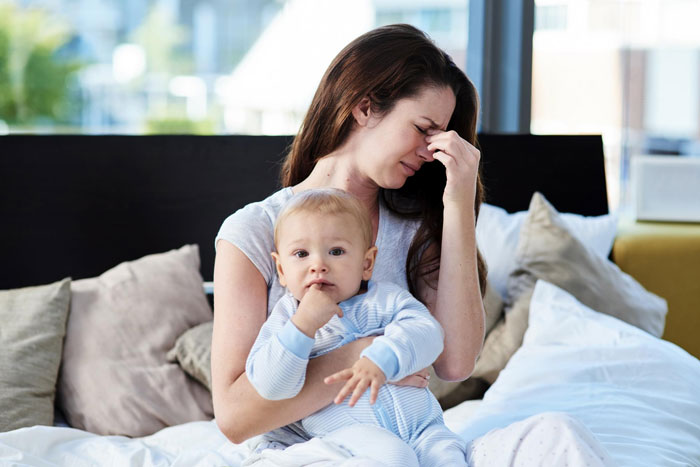 Tired mother holding newborn baby in pajamas on bed, showing emotional strain, related to mil keeps kissing newborn. Tired mother holding newborn baby in pajamas on bed, showing emotional strain, related to mil keeps kissing newborn.