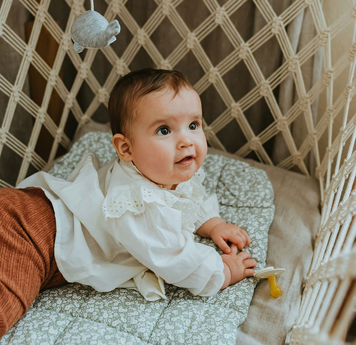 Mom takes her baby; infant lying in woven crib looking up, wearing white blouse with pacifier beside her Mom takes her baby; infant lying in woven crib looking up, wearing white blouse with pacifier beside her