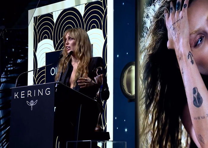 Miley Cyrus speaking at Kering event with bold tattoos visible, embodying strength and pushing back at photographers. Miley Cyrus speaking at Kering event with bold tattoos visible, embodying strength and pushing back at photographers.