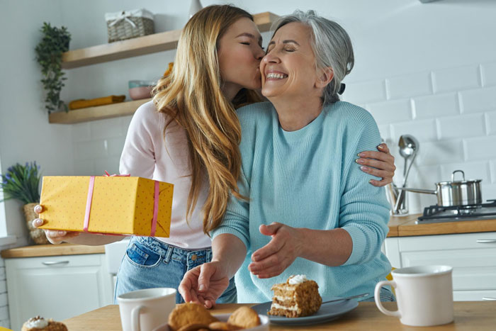 Young woman kissing happy mother holding birthday gift in kitchen, highlighting bros too broke to pitch in scenario.